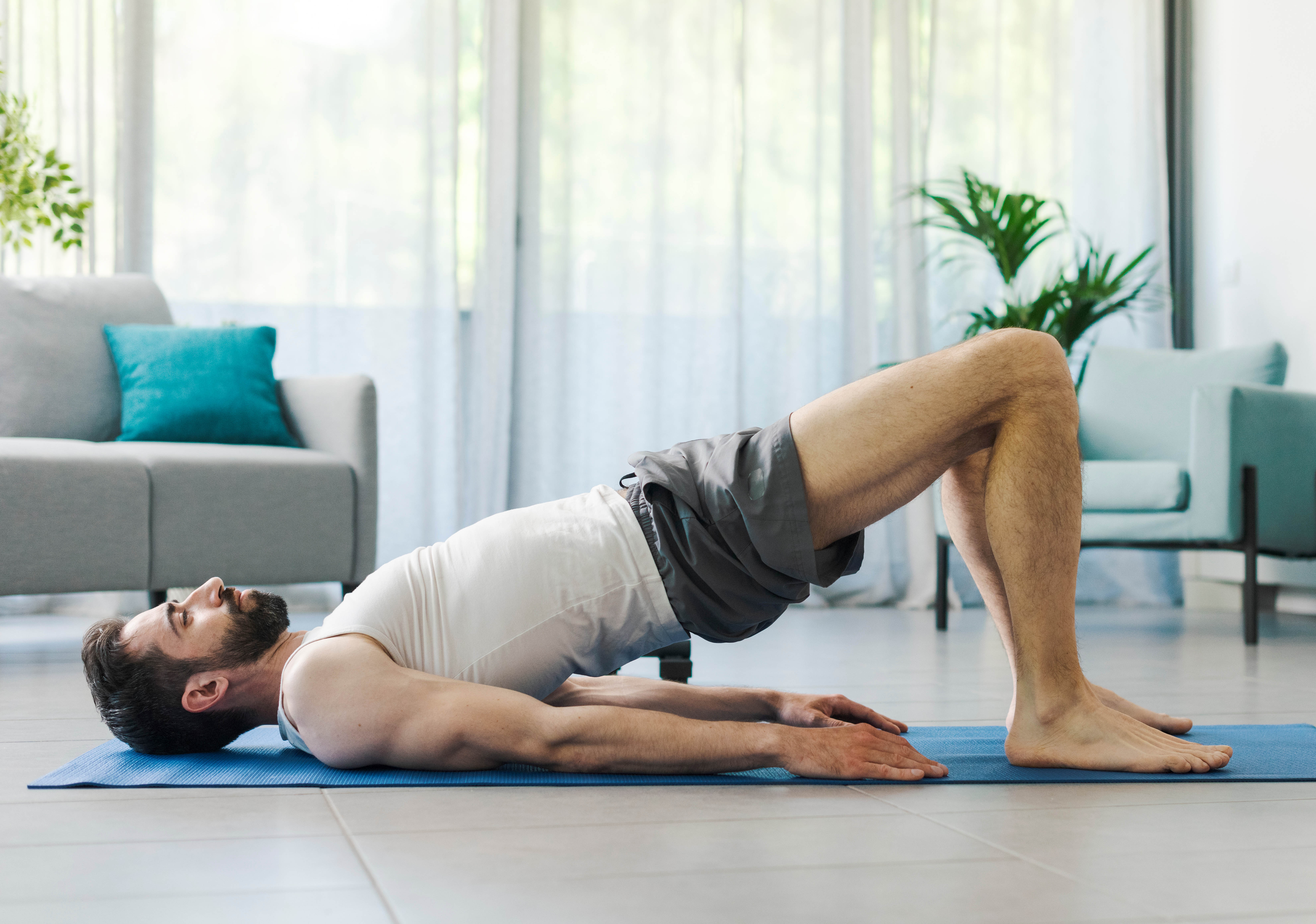 Athletic man in a bridge pose on a yoga mat in his living room; couch, chair, and plants in the background